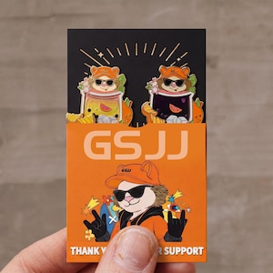Op de afbeelding: Een oranje en zwarte kaart met twee emaille pins met beer karakters met zonnebrillen en hoeden, die drankjes vasthouden. De kaart heeft ook een beer karakter met een hoed en zonnebril. De tekst "GSJJ" en "THANK YOU FOR SUPPORT" zijn zichtbaar.