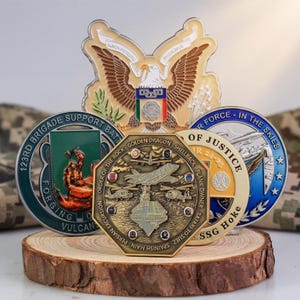 Puede incluir: Una colección de monedas de desafío militares exhibidas sobre una base de madera. Las monedas presentan varios diseños, incluyendo un águila, un barco y texto como "123rd Brigade Support Bat" y "Order of the Rock". Las monedas son en tonos dorados, plateados, verdes y azules.