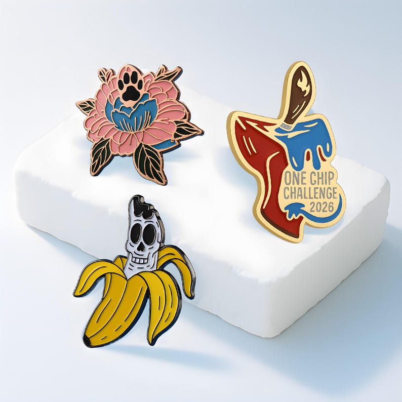 May include: Three enamel pins featuring distinct designs: a pink and blue flower with a black paw print center, a red and blue chip shape with a flame icon and the text ONE CHIP CHALLENGE 2026, and a yellow banana peel revealing a white skull.
