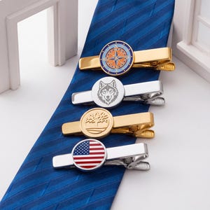 May include: Four tie clips with different designs are displayed on a blue striped tie. The clips include a gold compass design, a silver wolf head, a gold tree design, and a silver clip with an American flag.