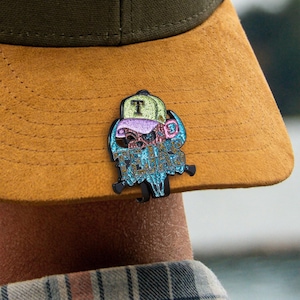 May include: A colourful enamel pin depicting a skull wearing a baseball cap. The cap is green with a black "T" on the front. The skull has purple, blue, and pink accents, with the words "Texas Teguards" in blue. The pin is attached to a tan hat.