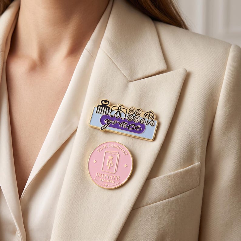 May include: A rectangular enamel pin featuring a light blue background, a purple oval with the word grace in cursive, and four black symbols above. Below it is a circular pink enamel pin with the text Pink Passport and Initiate Stamped.
