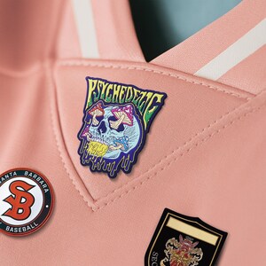 May include: Close-up of a pink athletic shirt with white stripes and patches. The patches include a skull with mushrooms and the word "PSYCHEDELIC", a Santa Barbara baseball logo, and a black and gold emblem.