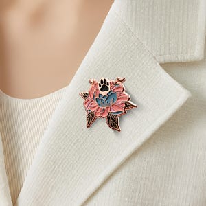 May include: A floral-shaped brooch with a paw print in the center. The brooch features pink, blue, and black enamel with gold-colored metal edges. The brooch is pinned to a white blazer.