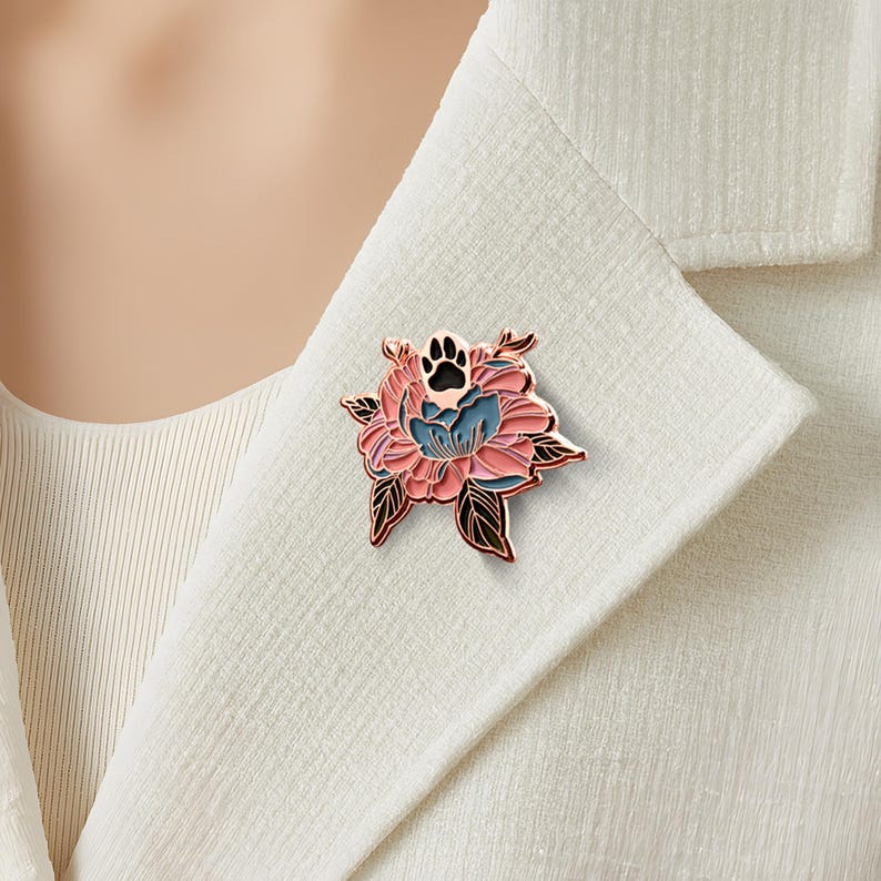May include: A metal enamel pin featuring a stylized pink and blue flower with a black paw print at the center. The design is outlined in a rose gold tone, showcasing layered petals and dark leaves, attached to the lapel of a white textured blazer.