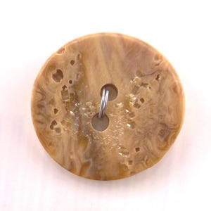 May include: A large, round button with a marbled, tan and brown pattern. The button has two holes and a metal shank. The button has a unique, textured surface with small holes and imperfections.