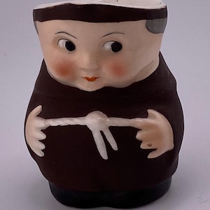 Puede incluir: Figura de cerámica de un monje con túnica marrón y cuello blanco. La cara está pintada con mejillas rosadas, ojos negros y una boca roja pequeña. La figura tiene una base negra.