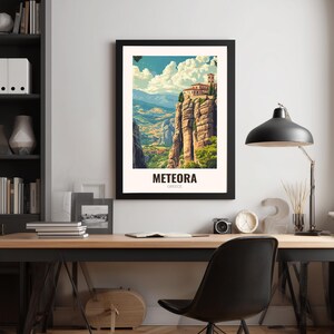 May include: A framed art print of Meteora, Greece. The image shows a monastery perched on a rock formation, with a valley and mountains in the background. The print is in a black frame, with the word "METEORA" printed at the bottom.