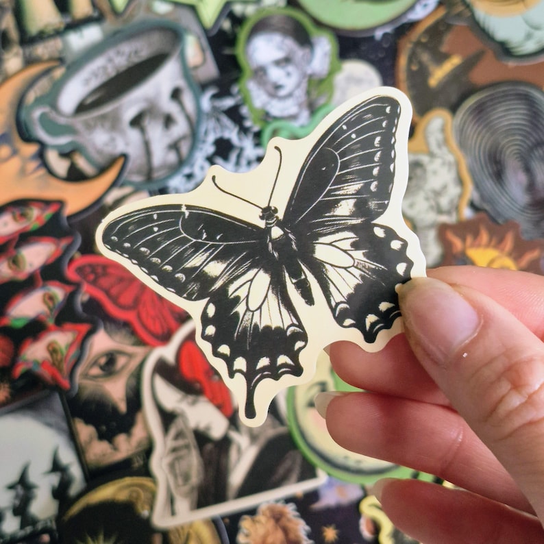 May include: A die-cut sticker featuring a black and cream butterfly illustration with intricate wing patterns and detailed veining. The butterfly is depicted in a high-contrast graphic style with a solid black body and wings against a cream background.