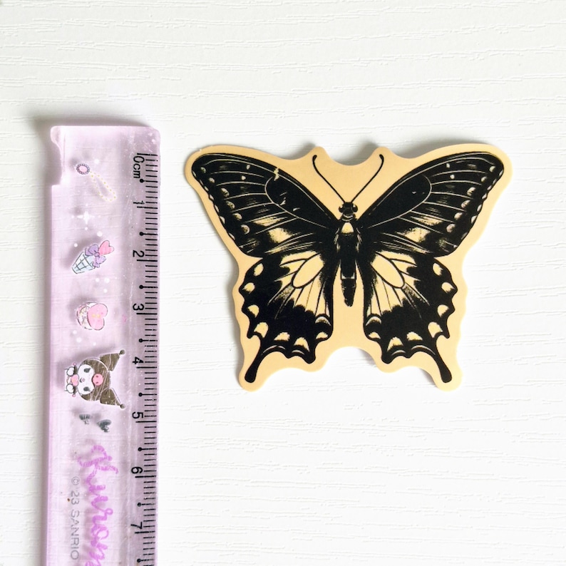 May include: A die-cut sticker featuring a black and cream butterfly illustration. The sticker is positioned next to a translucent purple ruler that shows the butterfly has a wingspan of approximately 3 inches and a height of 2.5 inches.