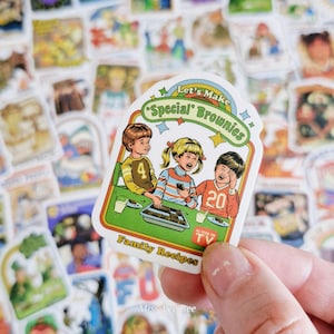 May include: A die-cut sticker featuring a retro illustration of three children at a table with a tray of brownies. The text reads Let's Make Special Brownies and Family Recipes, with an As Seen On TV logo. The design uses green, orange, and blue.