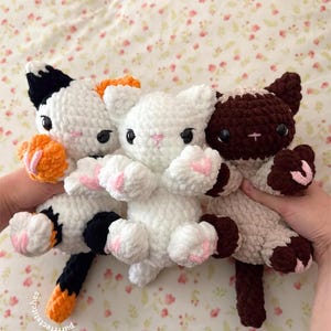 May include: Three handmade crochet cat plushies in white, calico, and brown. The white cat has pink paw pads, while the calico cat has orange and black accents. The brown cat has brown and cream accents. The plushies are soft and cuddly.