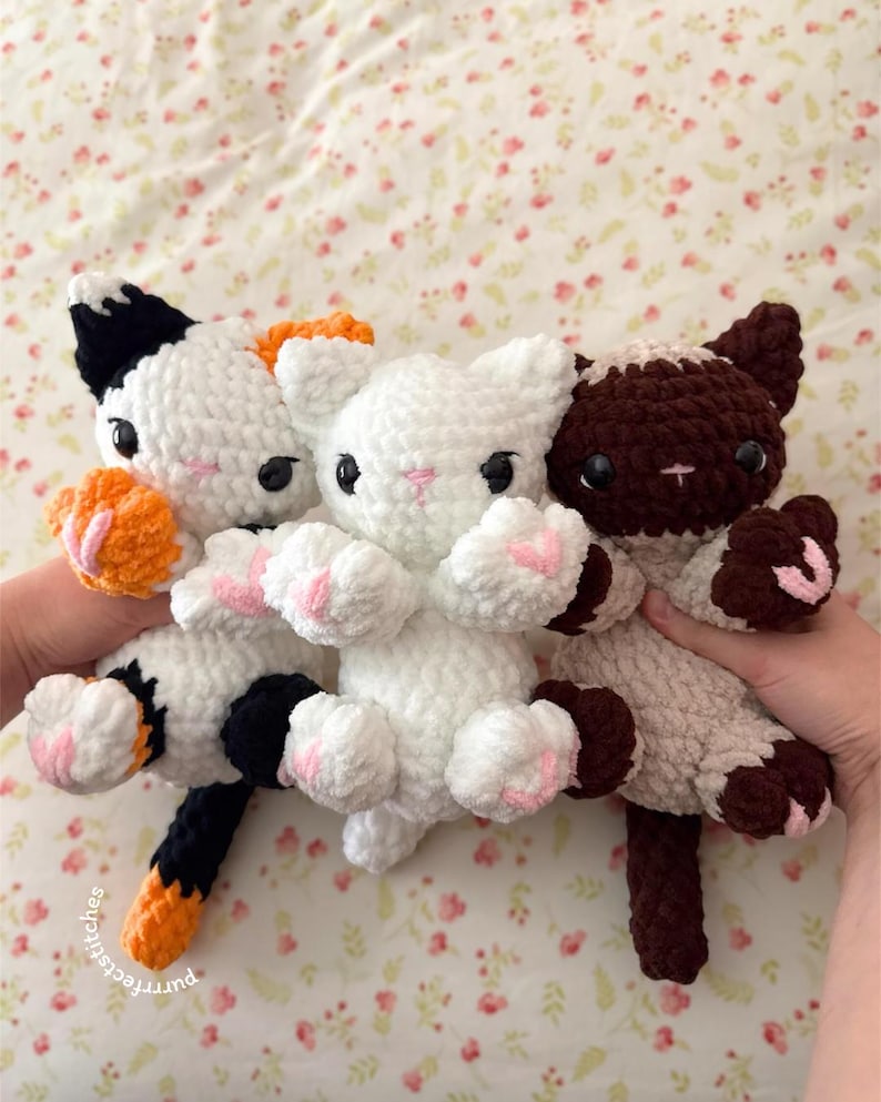 May include: Three handmade crochet cat plushies in white, calico, and brown. The white cat has pink paw pads, while the calico cat has orange and black accents. The brown cat has brown and cream accents. The plushies are soft and cuddly.