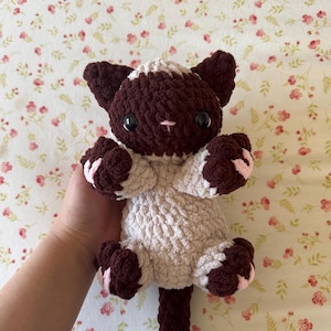 May include: A handmade crochet cat plushie in shades of brown and cream. The cat has large black eyes, a pink nose, and pink accents on its paws. The plush toy is soft and cuddly, perfect for children or cat lovers.