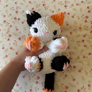 May include: A handmade crochet cat doll in white, black, and orange. The cat has black eyes, pink accents on its paws, and orange ears. The cat is being held up against a floral background.