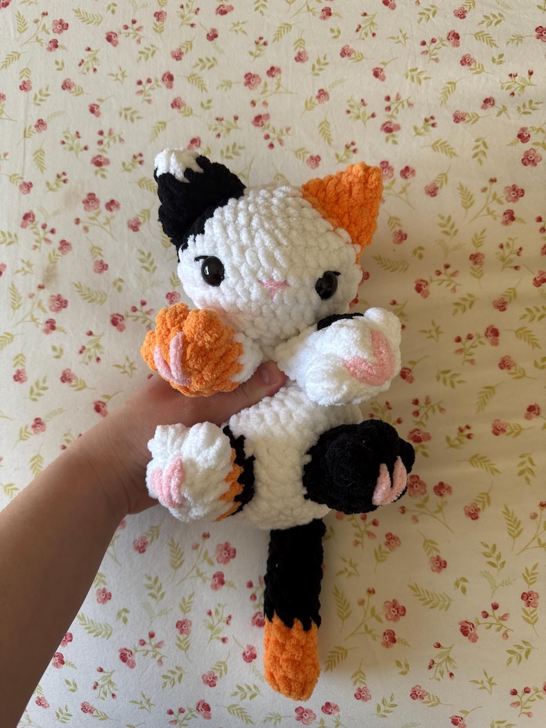 May include: A handmade crochet cat doll in white, black, and orange. The cat has black eyes, pink accents on its paws, and orange ears. The cat is being held up against a floral background.