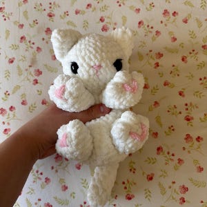 May include: A handmade white crochet cat plushie with black eyes and pink accents on the paws and nose. The soft, fluffy toy is designed in a lying down position, perfect for a child's toy or nursery decor.