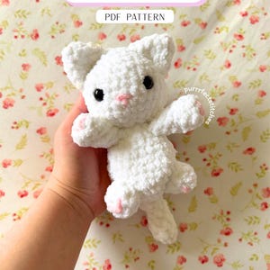 May include: A handmade white crochet kitten with black eyes and pink accents on the nose and paws. The text "Baby Kitty" and "PDF PATTERN" are displayed on a banner above the kitten. The kitten is held against a floral background.