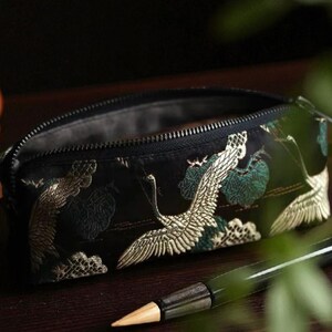 以下が含まれることがあります： 黒色のペンケース。白い鶴と緑の葉が刺繍されています。ペンケースは、暗い木製の表面に置かれ、背景にはペンと小さな茶色の瓶があります。