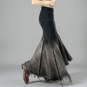 以下が含まれることがあります： フィッシュテールのデザインが施された黒色のデニムスカート。スカートは、黒からグレーに変化する、色あせた、使い古されたグラデーション効果が特徴です。裾はほつれたエッジになっています。茶色の革靴と合わせています。