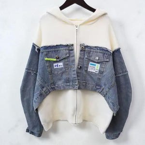 以下が含まれることがあります： クリーム色のボディと袖、青色のデニムフロントが付いたフード付きデニムジャケット。 ジャケットにはフロントポケットが2つとジッパー開閉が付いています。 ポケットにはパッチが見えます。 ジャケットは黒いハンガーに吊るされています。