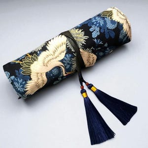 May include: A rolled-up fabric case with a black background, adorned with white cranes and blue floral patterns. The case is secured with a black leather strap and features two navy blue tassels with gold accents.