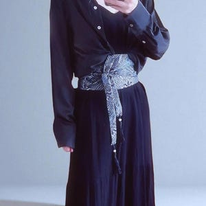 以下が含まれることがあります： 長袖ボタンダウンシャツ、お揃いのスカート、装飾的な模様のサッシュが特徴の濃いネイビーブルーの服装。 サッシュはウエストに結ばれ、詳細なデザインが施されています。 この服装は、さまざまな機会に適しています。