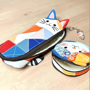 Puede incluir: Un estuche con forma de gato y un monedero redondo con un llavero a juego. El estuche es largo y rectangular, con un diseño de patchwork en rojo, naranja, azul y blanco. El monedero tiene un diseño similar.