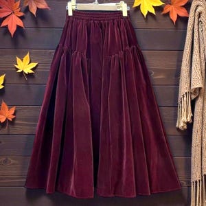 以下が含まれることがあります： ハンガーに吊るされた、秋の葉で飾られた木製の背景に、長いバーガンディ色のベルベットスカート。右側にはフリンジ付きのベージュのスカーフが掛かっています。スカートはギャザーウエストで、流れるようなフレアの形をしています。