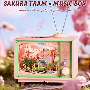 以下が含まれることがあります： 「SAKURA TRAM x MUSIC BOX」と「A Journey Through Springtime Melodies」の文字が入った、レトロテレビ型のピンク色のオルゴール。画面には、緑色の路面電車と桜の3Dジオラマが表示されています。