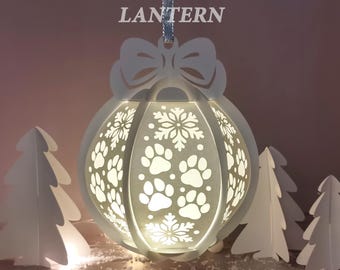 Peut inclure: Une lanterne boule de Noël blanche avec des découpes de pattes de chien et de flocons de neige. La lanterne est ornée d'un nœud en haut et suspendue par un ruban argenté. Le texte "CHRISTMAS BALL LANTERN" est en haut. Des sapins en papier blanc entourent la lanterne.
