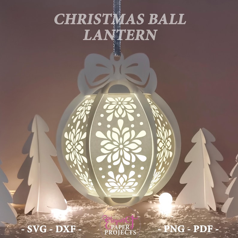 May include: A white Christmas ball lantern with intricate floral cutouts, suspended by a silver ribbon. The lantern is surrounded by white paper Christmas trees and a soft, glowing light. The text "CHRISTMAS BALL LANTERN" is at the top.
