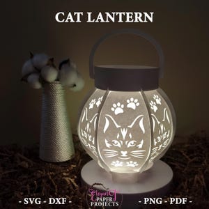 Könnte beinhalten: Eine weiße Katzenlaterne mit Griff, mit Katzengesichtern und Pfotenabdrücken, die in die Seiten geschnitten sind. Der Text "CAT LANTERN" steht oben. Die Laterne steht auf einem weißen Sockel.