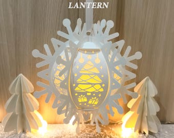 Peut inclure: Une lanterne flocon de neige blanche suspendue avec un intérieur lumineux, présentant un motif d'arbre de Noël. La lanterne est entourée de découpes de flocons de neige et de deux arbres de Noël en papier. Le texte "HANGING SNOWFLAKE LANTERN" est en haut.