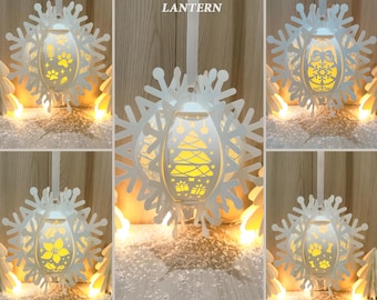 May include: White hanging snowflake lantern with a central oval-shaped light. The lantern features cut-out designs of a Christmas tree, paw prints, and gifts. The text "HANGING SNOWFLAKE LANTERN" is at the top. The bottom text reads "- SVG - DXF -" and "- PNG - PDF -".