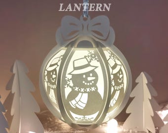 Peut inclure: Une lanterne boule de Noël blanche avec un motif de bonhomme de neige. Le texte "CHRISTMAS BALL LANTERN" est en haut. La lanterne a un nœud en haut et est suspendue à un ruban argenté. Le texte "SVG - DXF - PNG - PDF" est en bas.