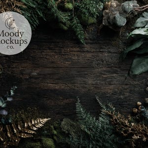 May include: Dark, textured wooden surface with a border of green foliage, moss, and dried plants. The image features the text "Moody Mockups co." in a circular logo, creating a natural, rustic aesthetic, suitable for product displays.