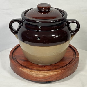 May include: A two-toned ceramic crock pot with a lid and two handles, sitting on a round wooden trivet. The crock pot is brown and tan, and the lid has a small knob. The trivet is made of dark wood.