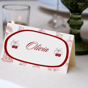 May include: A folded place card with a white background and a red oval border. The name "Olivia" is written in elegant script within the oval. The card is decorated with cherry and cake illustrations.