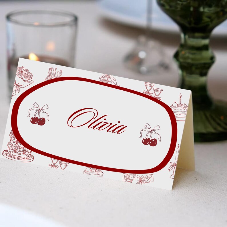 May include: A folded place card with a white background and a red oval border. The name "Olivia" is written in elegant script within the oval. The card is decorated with cherry and cake illustrations.