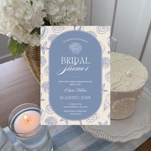 May include: A bridal shower invitation with a blue and cream seashell design. The invitation includes the text "Bridal Shower", the bride's name, and the date. A white cake with seashell decorations and a lit candle are also visible.