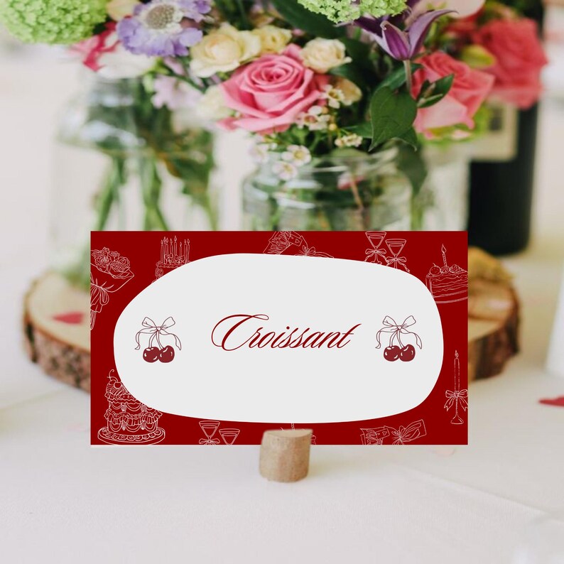 May include: A burgundy place card with the word "Croissant" in elegant script. The card features a white oval with illustrations of cakes, cherries, and flowers. A cork base supports the card, with a blurred floral arrangement in the background.
