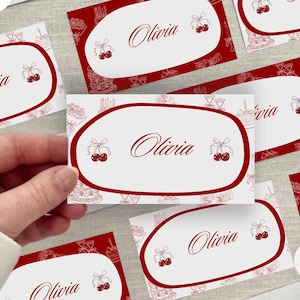 May include: White place cards with a red border and background. The cards feature the name "Olivia" in elegant script, along with cherry illustrations. The background has a pattern of cakes, candles, and glasses.
