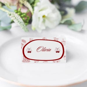 May include: A white place card with a red oval border and the name "Olivia" in cursive. The card is decorated with red illustrations of cakes, cherries, and other celebratory elements. The card is on a white plate.