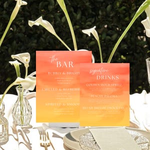 May include: Two rectangular bar signs with a watercolor gradient of orange and yellow. The signs read "The Bar" and "Signature Drinks" in elegant script. The signs list drink options like "Golden Hour Spritz" and "Peachy Paloma". White calla lilies and glassware are in the foreground.