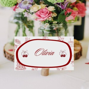 May include: A white place card with a burgundy border and the name "Olivia" in elegant script. The card features decorative illustrations of cakes, cherries, and glasses. It is held by a small wooden stand, with a blurred floral arrangement in the background.