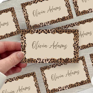 May include: A hand holding a beige card with the name "Olivia Adams" in elegant script. The card has a scalloped edge and a leopard print border. Other similar cards are visible in the background.