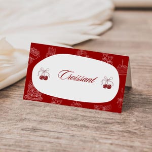May include: A red place card with a white oval in the center. The word "Croissant" is written in a cursive font. The card is decorated with white line drawings of cakes, cherries, and other pastries.