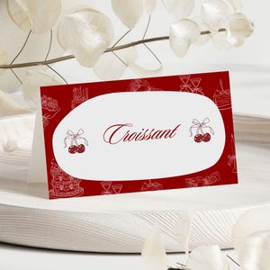 May include: A red place card with a white oval in the center. The word "Croissant" is written in a cursive font. The card is decorated with white line drawings of cakes, cherries, and other food items.