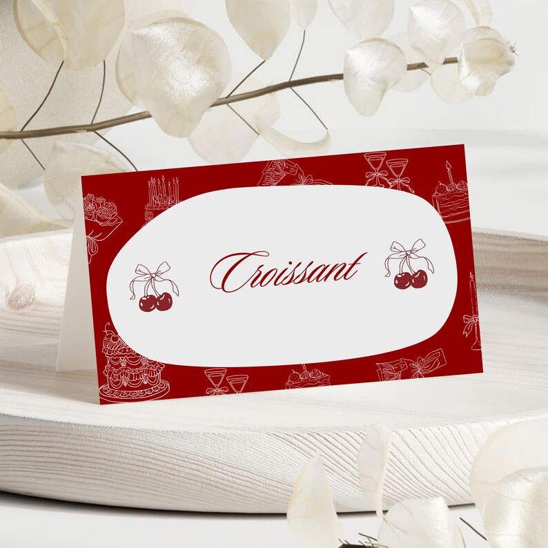 May include: A red place card with a white oval in the center. The word "Croissant" is written in a cursive font. The card is decorated with white line drawings of cakes, cherries, and other food items.